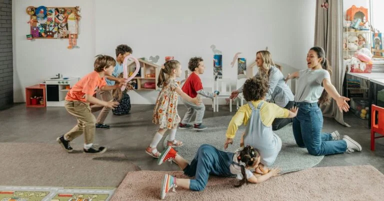 Children and two adults play together in a bright daycare room with toys, shelves, and a rug-filled floor.