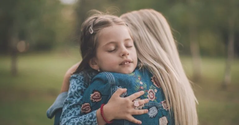 Young girl with eyes closed hugs a woman outdoors, both wearing denim jackets with floral embroidery or patterns on a park background.