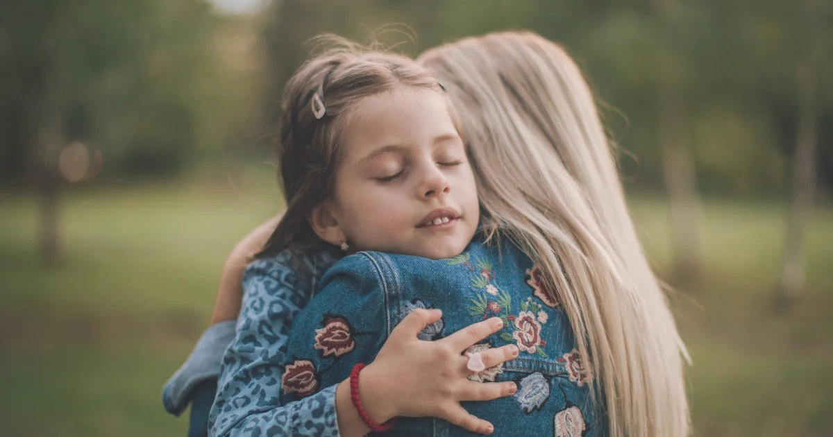 Young girl with eyes closed hugs a woman outdoors, both wearing denim jackets with floral embroidery or patterns on a park background.