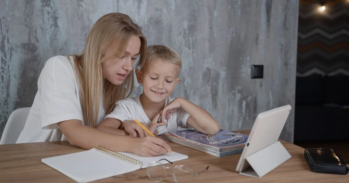 Mother helps her young son with homework at a wooden table, writing in a notebook with a pencil beside a tablet and books.