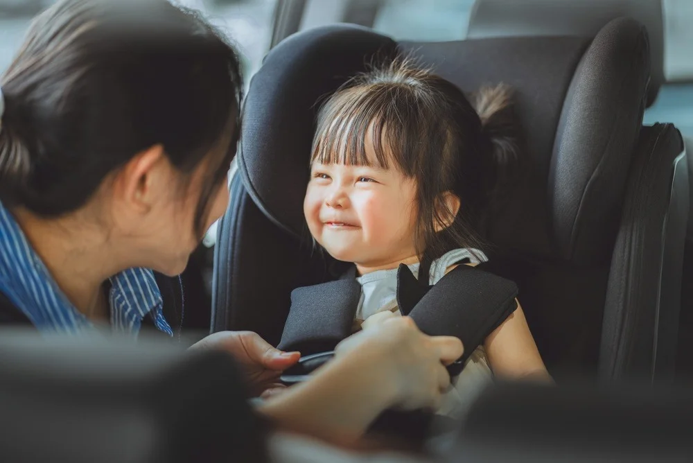 Young girl smiling while being buckled into a car seat by a caregiver in a car interior