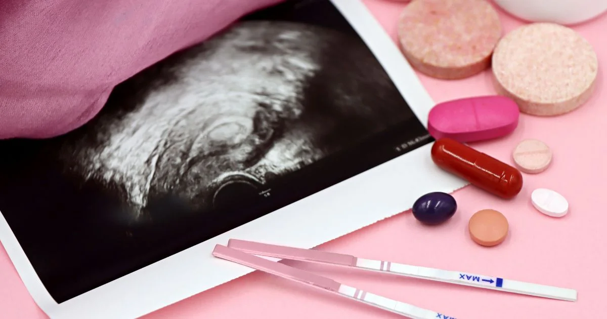 Ultrasound image on a tablet surrounded by pink pills and two pregnancy test sticks on a pink surface?